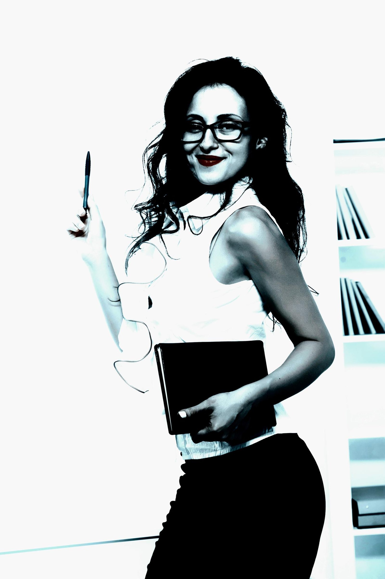 Playful yet professional woman with glasses holding a pen and notebook, smiling confidently in a brightly lit office.