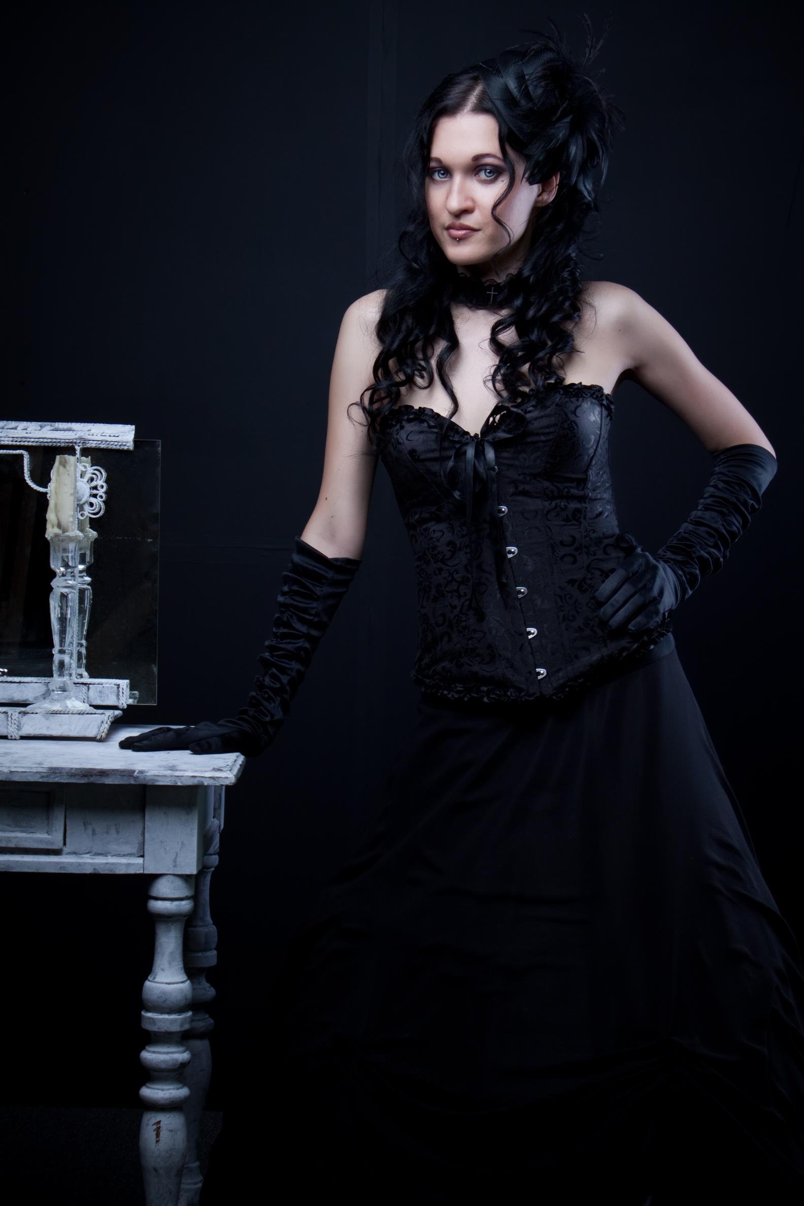 Elegant woman in a black corset dress with intricate lace details, standing beside a vintage table adorned with a candleholder.
