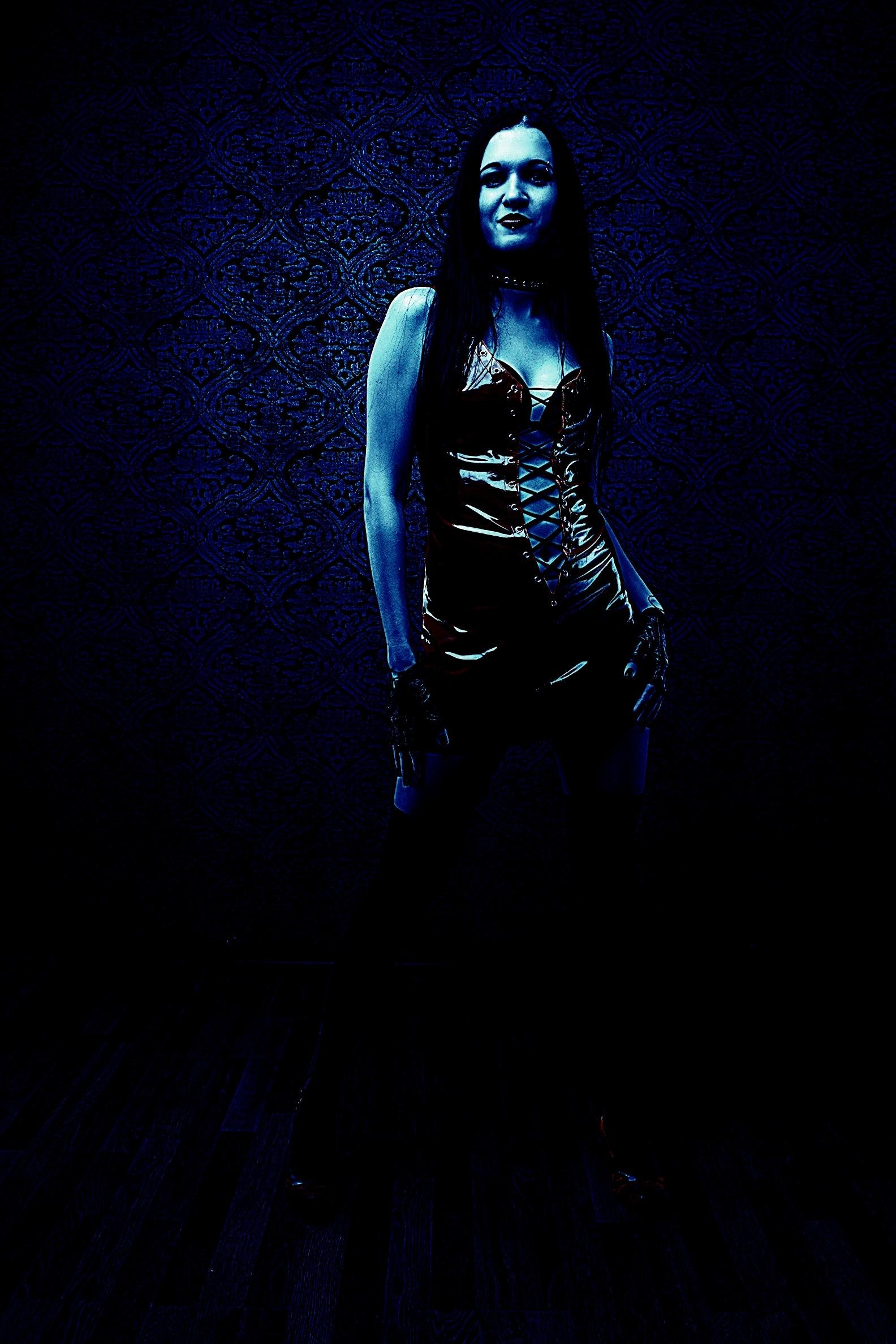 Dark and moody figure in a glossy black lace-up corset dress, illuminated with a blue-tinted light against a patterned background.