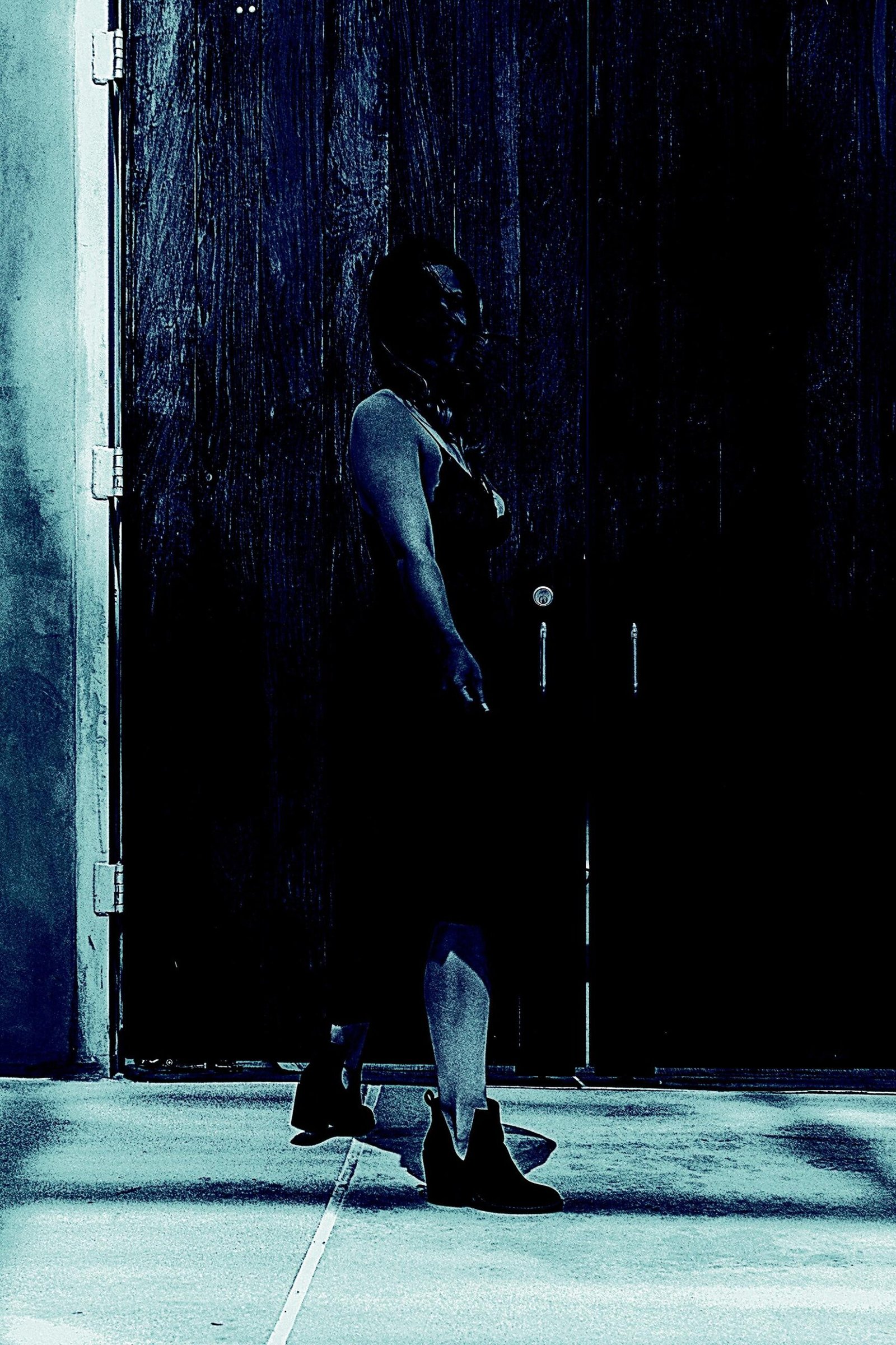 Silhouetted woman standing against a large, dark wooden door, partially lit with an eerie blue hue.