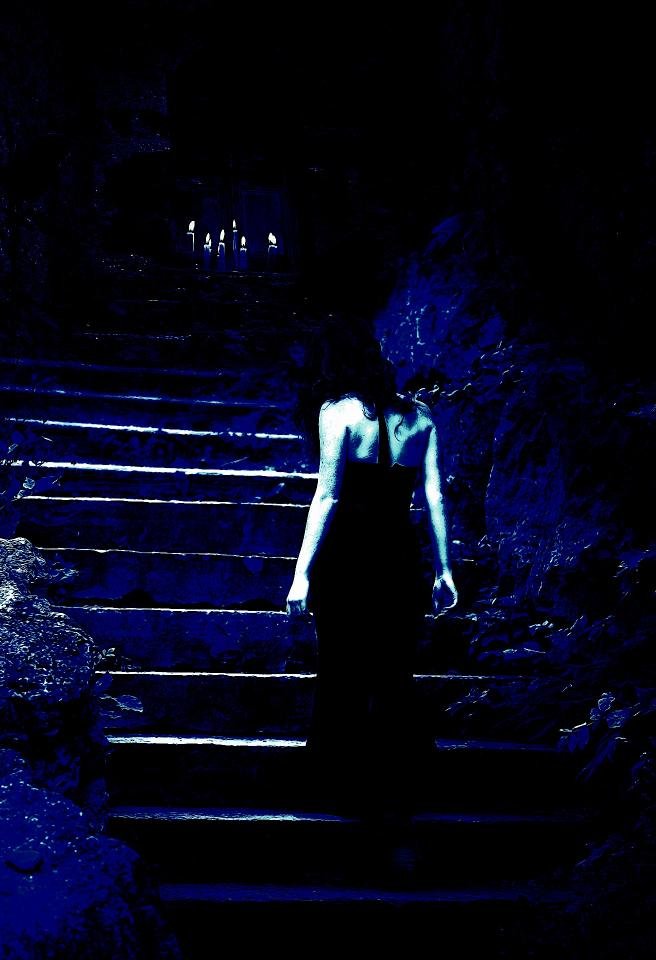 Back view of a mysterious woman in a black gown, descending a shadowy stone staircase lit by flickering candles in the distance.