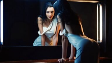 A glamorous woman in a white dress leaning against a mirror, illuminated by soft lighting that creates a seductive vibe.