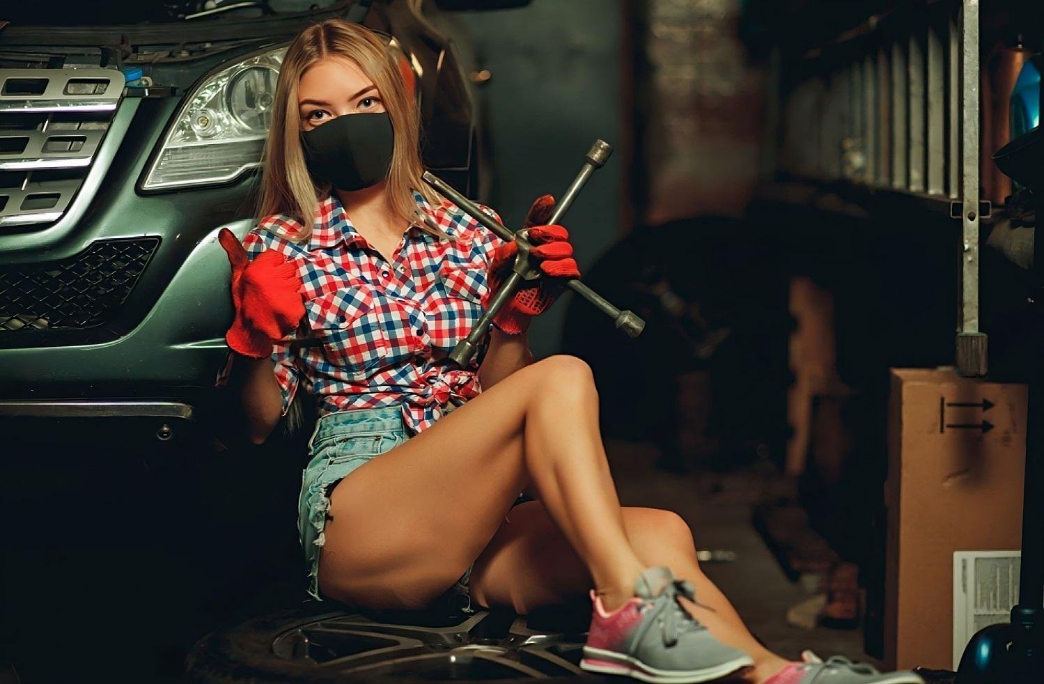 A blonde woman wearing a black face mask, plaid shirt, and denim shorts sits in a garage holding a tire iron, with a car behind her.