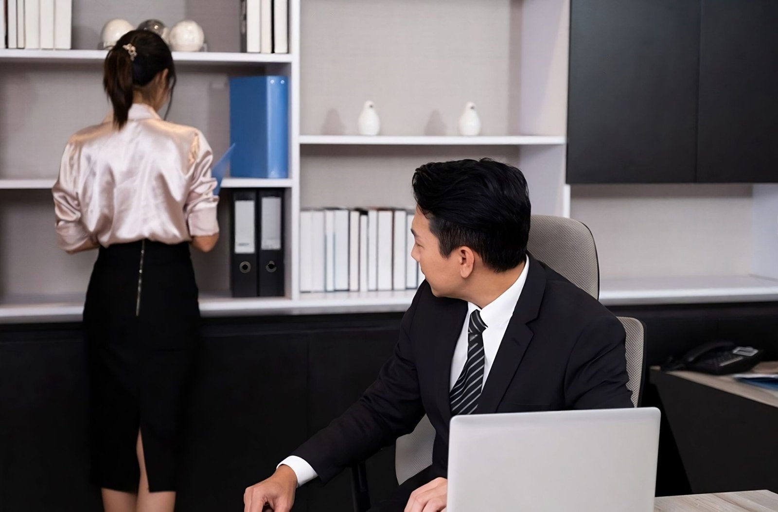 Businessman in an office chair looking at a female coworker in a silk blouse and tight skirt as she retrieves a file.