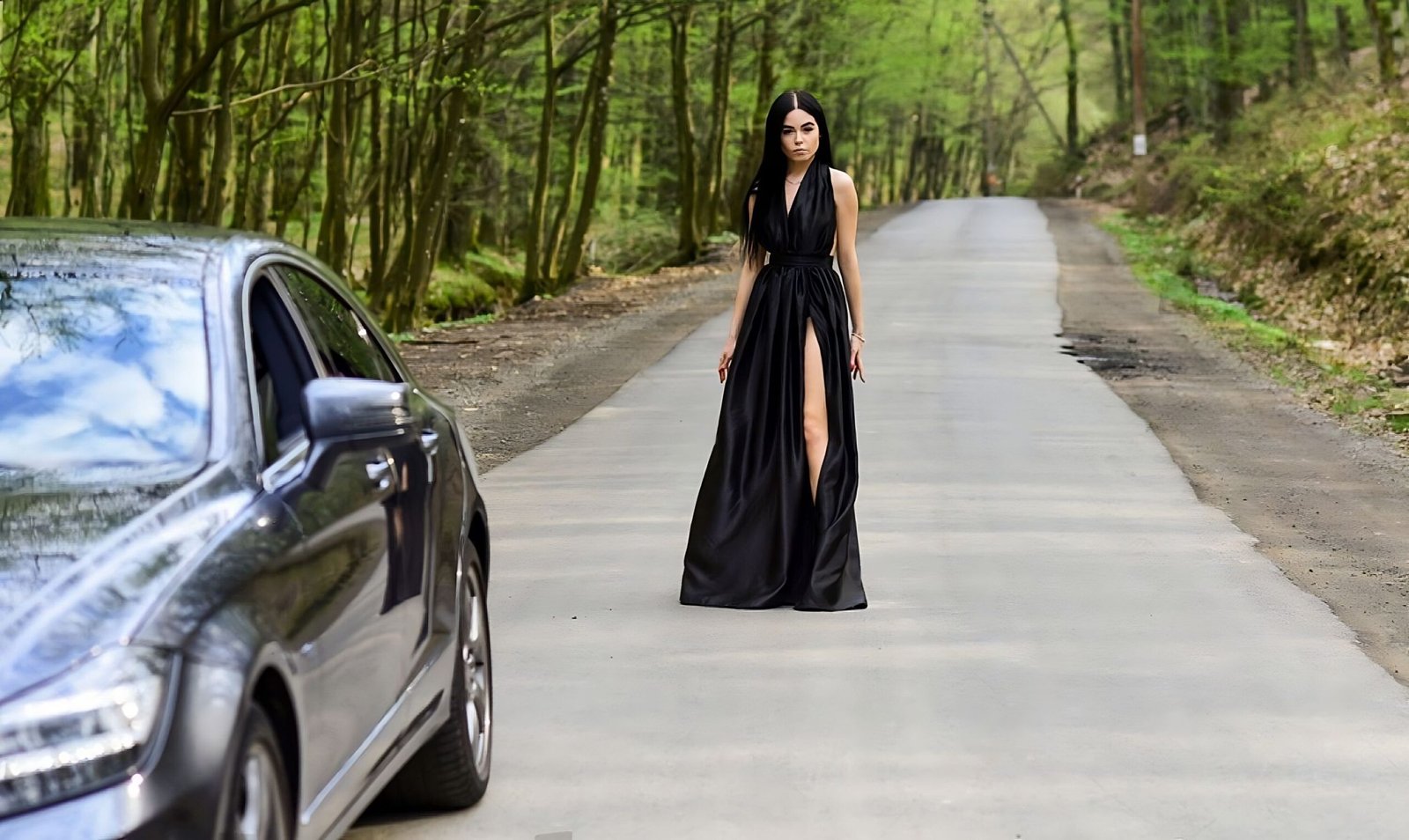 Gothic woman in a black dress with a thigh-high slit walking down a forest road towards a black car.