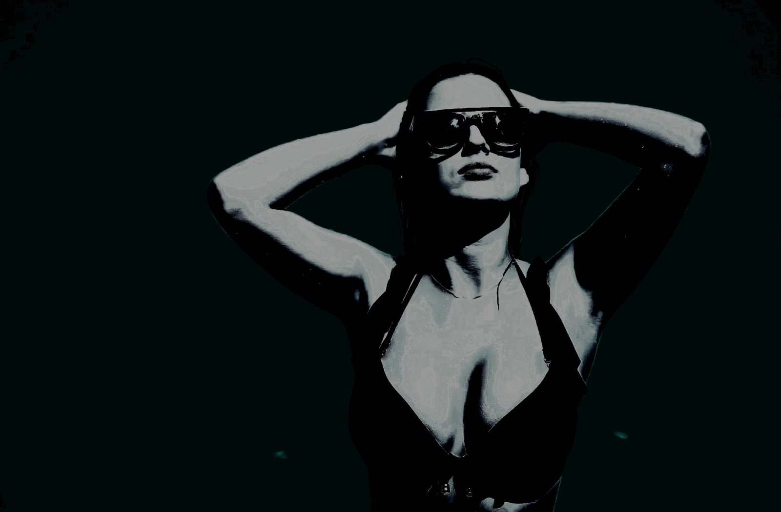 A woman in a black bikini and oversized sunglasses, confidently holding her hands behind her head against a shadowy background.