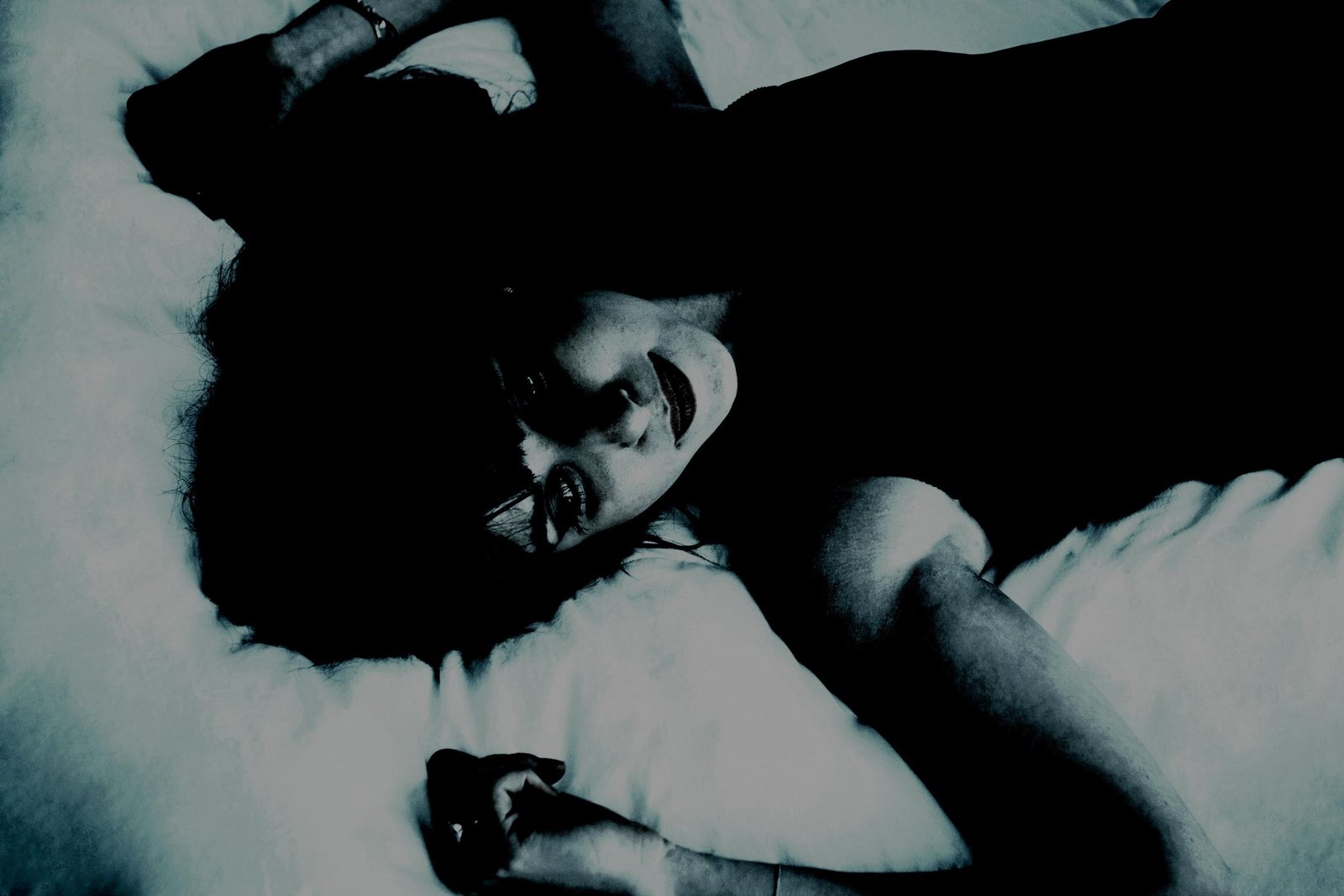 Mysterious woman in a dark dress lying on a bed, staring upward with a haunting expression.