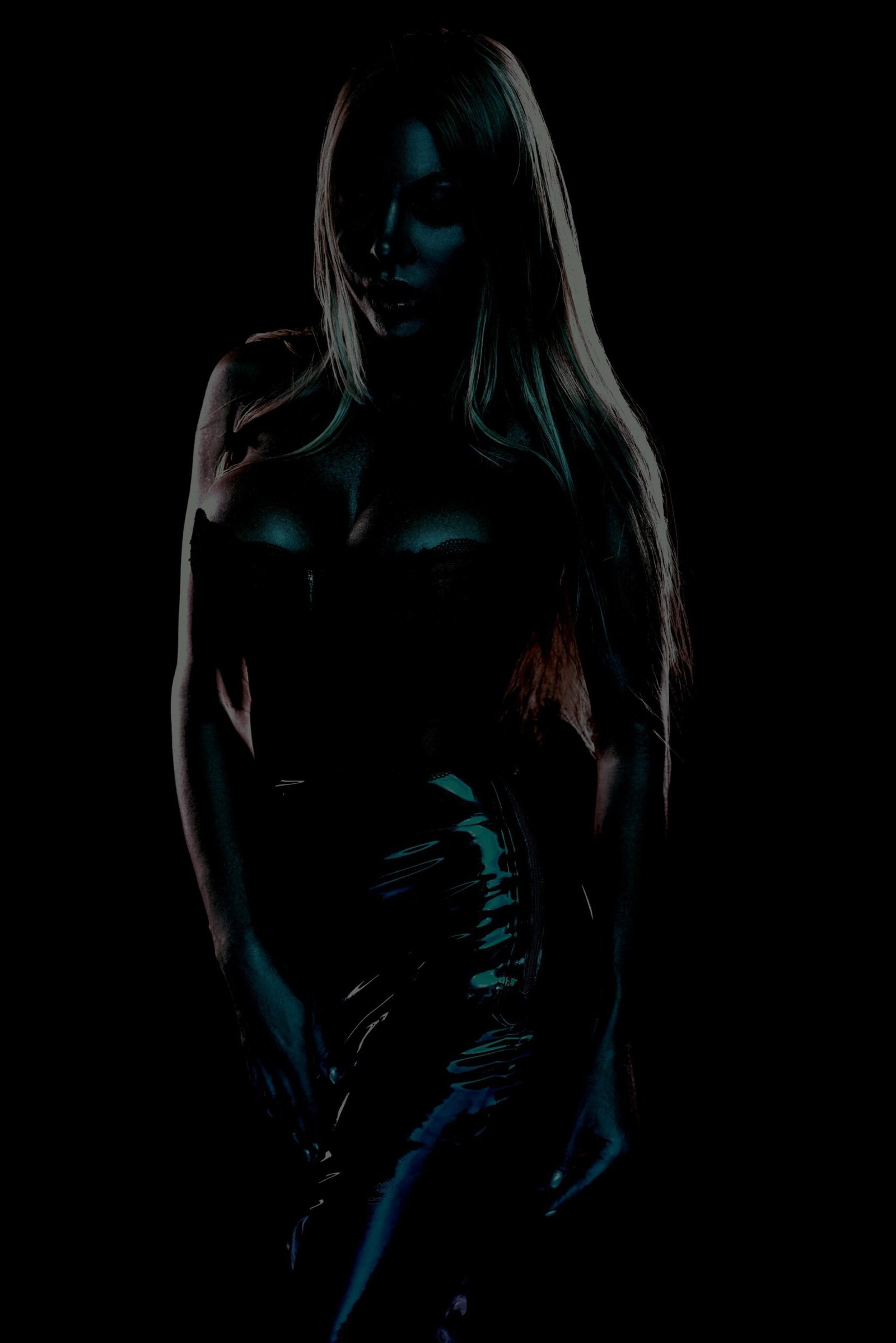 Sensual woman in a shiny black corset and latex skirt, illuminated softly in a dark setting.