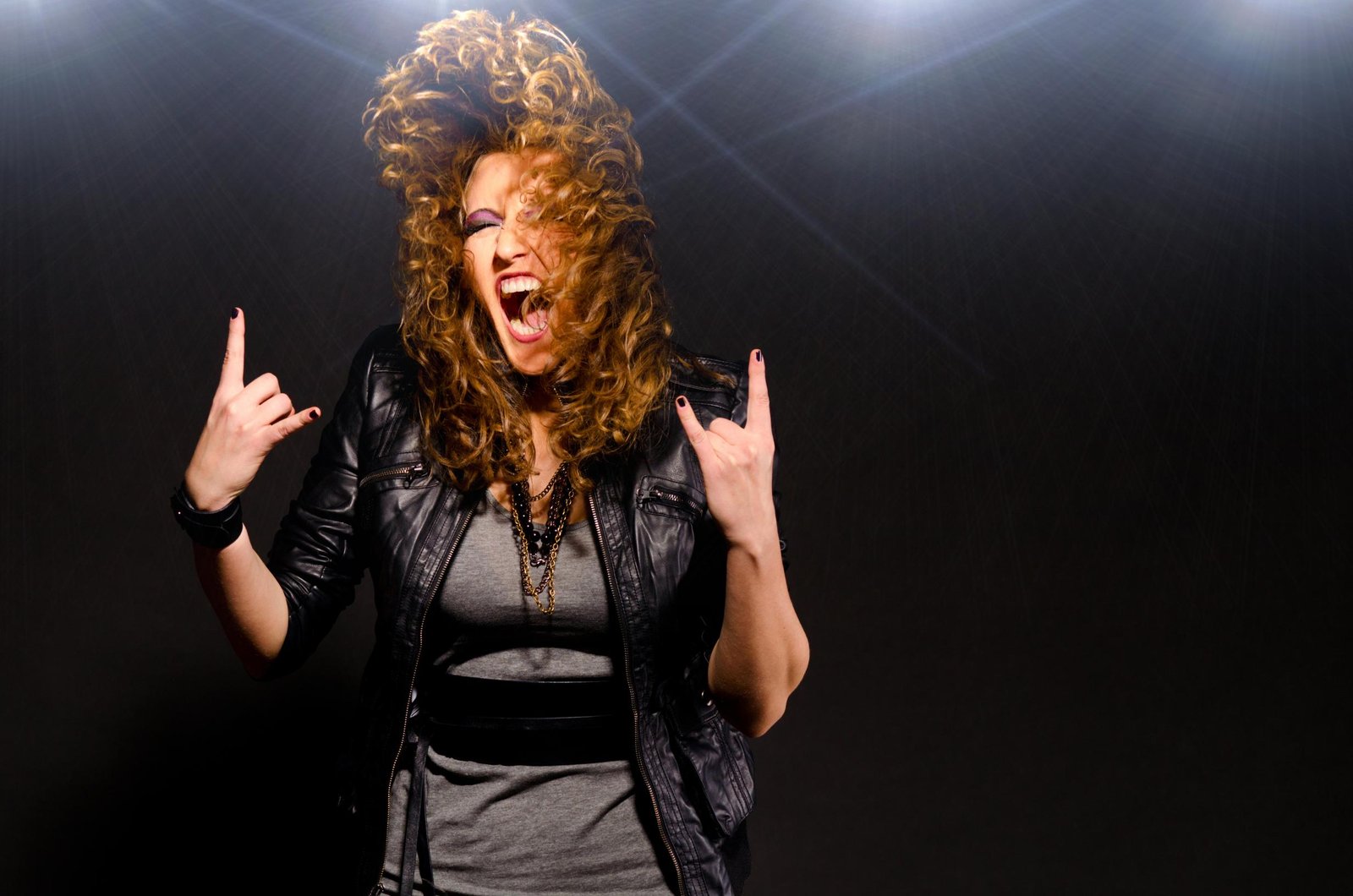 Curly-haired woman in black leather jacket and gray top, screaming with rock hand gesture on stage.
