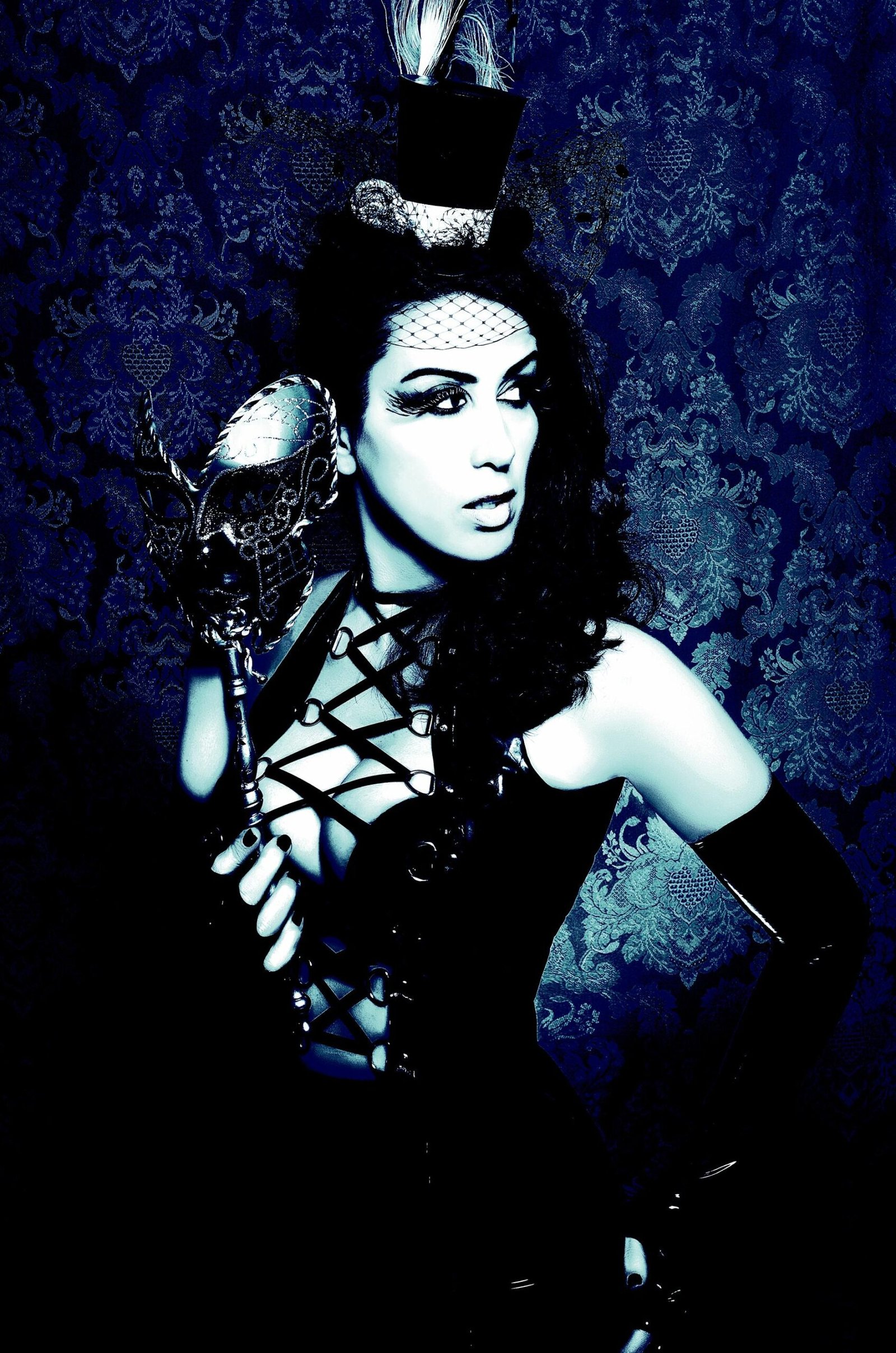 Mysterious gothic woman in a black corset top, holding a silver masquerade mask with a feathered mini top hat.