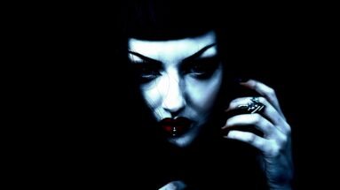 A gothic woman with dramatic makeup, dark arched eyebrows, red lips, and a lip piercing, gazing intensely in a moody, high-contrast portrait.