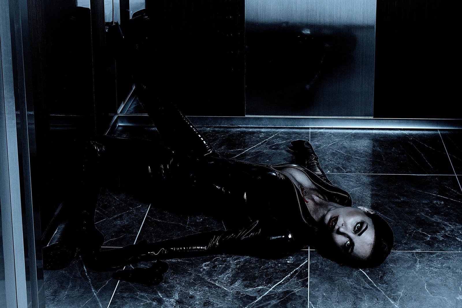Mysterious woman in a black latex bodysuit lying on a dark marble floor, staring intensely.