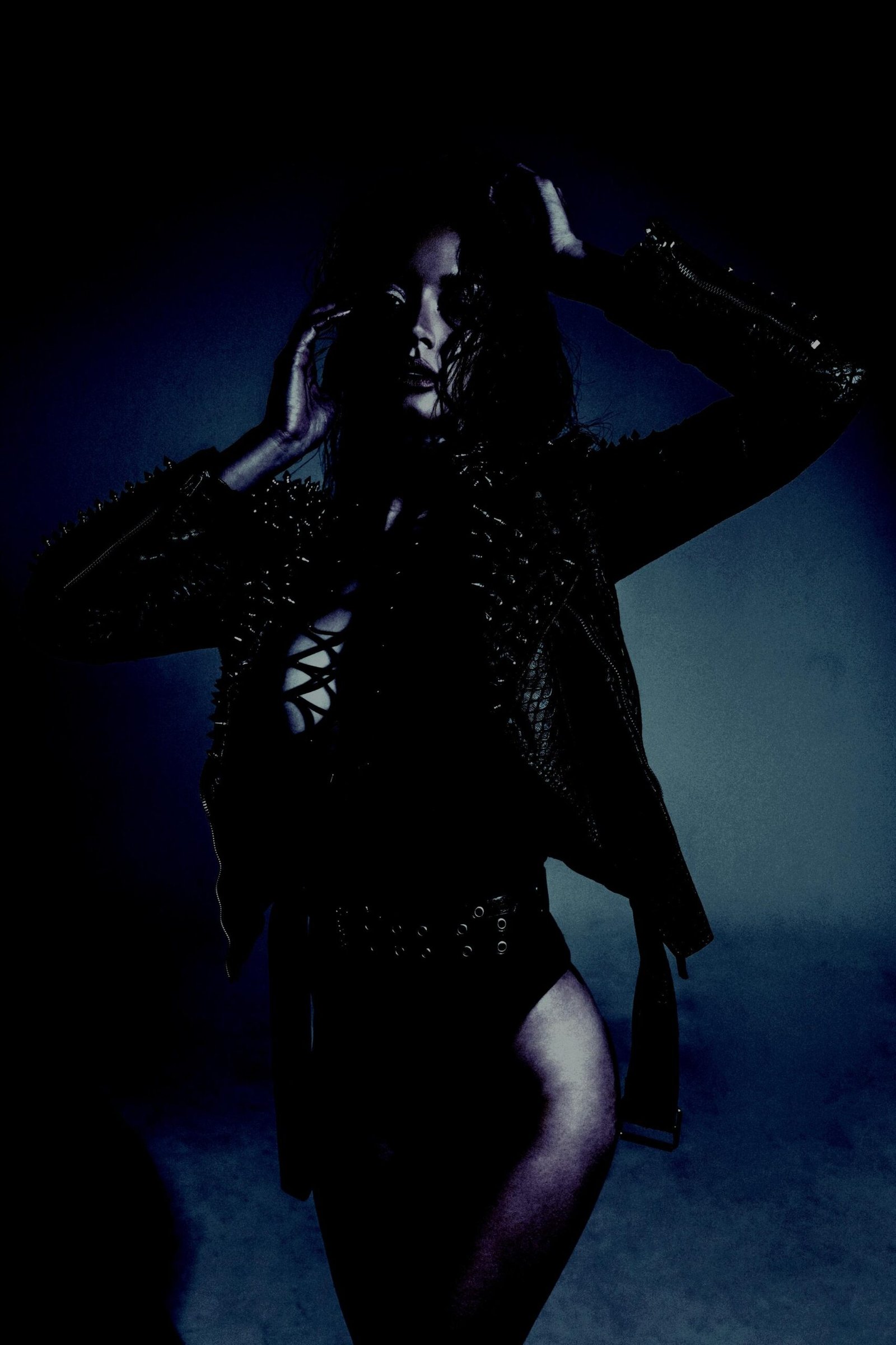 Bold woman in a spiked leather jacket and black bodysuit, striking a powerful pose in low lighting.