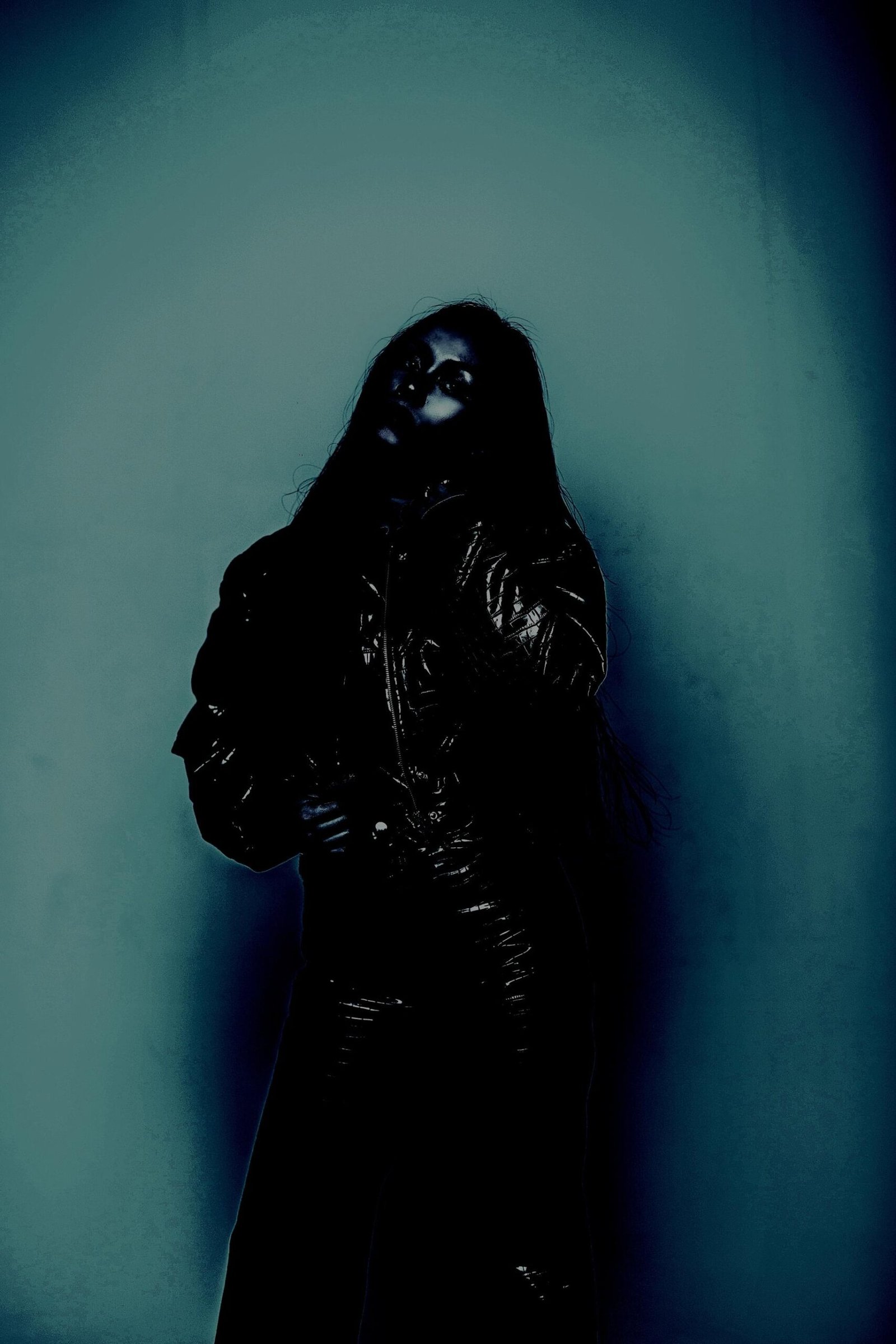 Woman in a black leather jacket and pants, standing against a teal-lit wall with a haunting gaze.