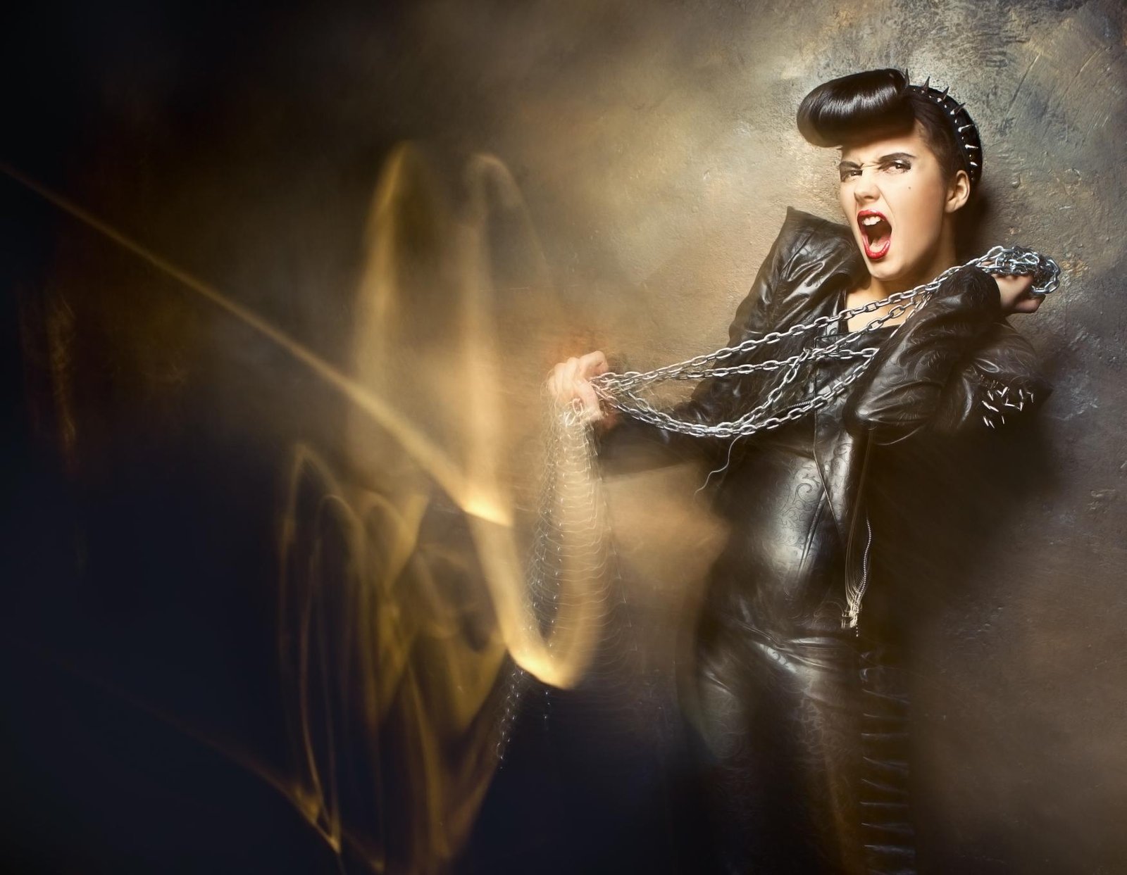 Woman in black leather catsuit with chains, spiked headband, dramatic makeup, and vintage pin-up hairstyle.
