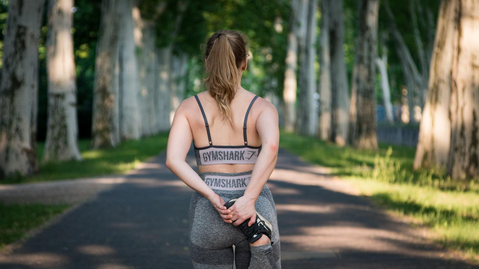 Woman in a Gymshark sports bra and leggings stretching her leg on a peaceful tree-lined path, embracing outdoor fitness recovery.