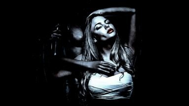 A woman with long hair and red lipstick posing sensually in low lighting, with a dark figure behind her.