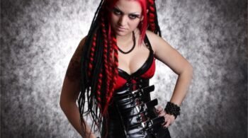 Gothic woman in a tight black leather corset with metal buckles, red lace bra, and black dreadlocks, striking a dominant pose.