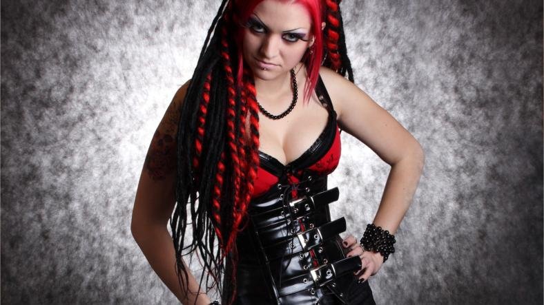 Gothic woman in a tight black leather corset with metal buckles, red lace bra, and black dreadlocks, striking a dominant pose.