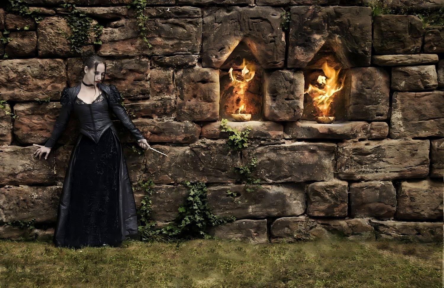 Witch in black corset dress standing by flaming alcoves in an old stone wall