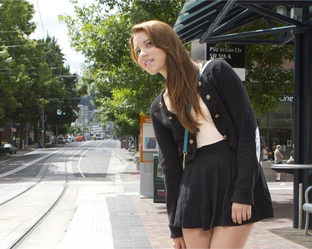 Woman waiting at a city bus stop in a pleated high-waisted black skirt and schoolgirl-inspired outfit