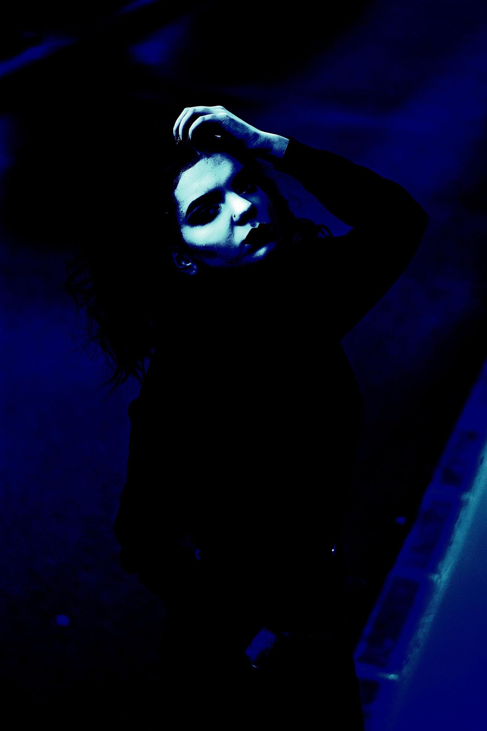 Goth woman in dramatic blue lighting holding her hair, wearing dark clothing and lip piercing