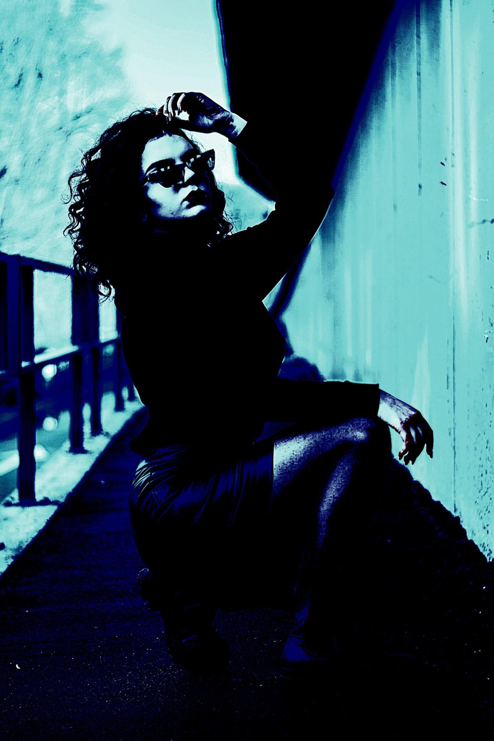 Curly-haired woman in sunglasses and a black outfit posing in an urban alleyway under harsh cyan lighting