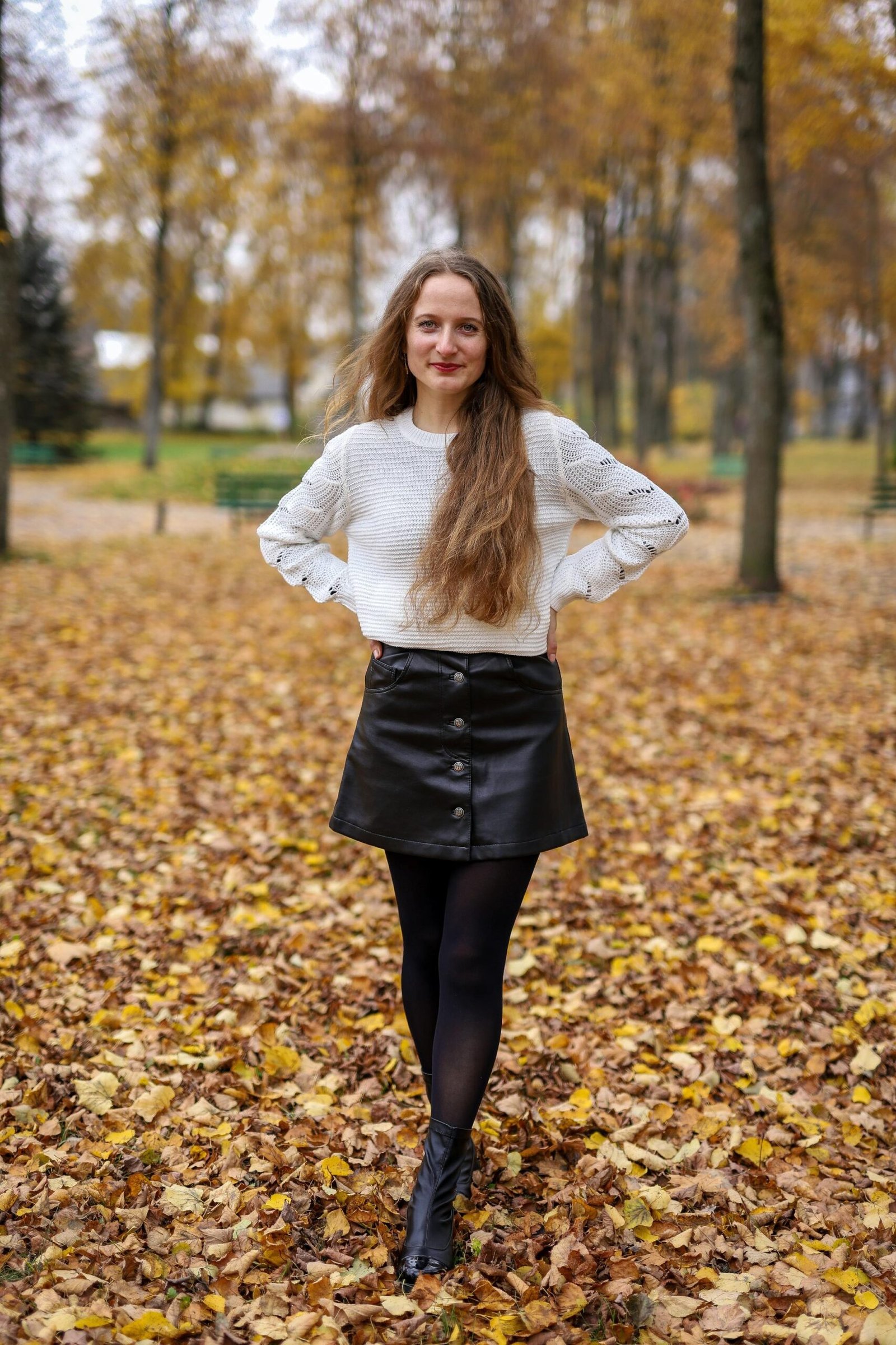 Woman standing in autumn leaves wearing a high-waisted black leather skirt and white sweater