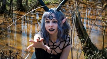 Gothic elf-like woman with horns and black lips reaching out in eerie swamp environment