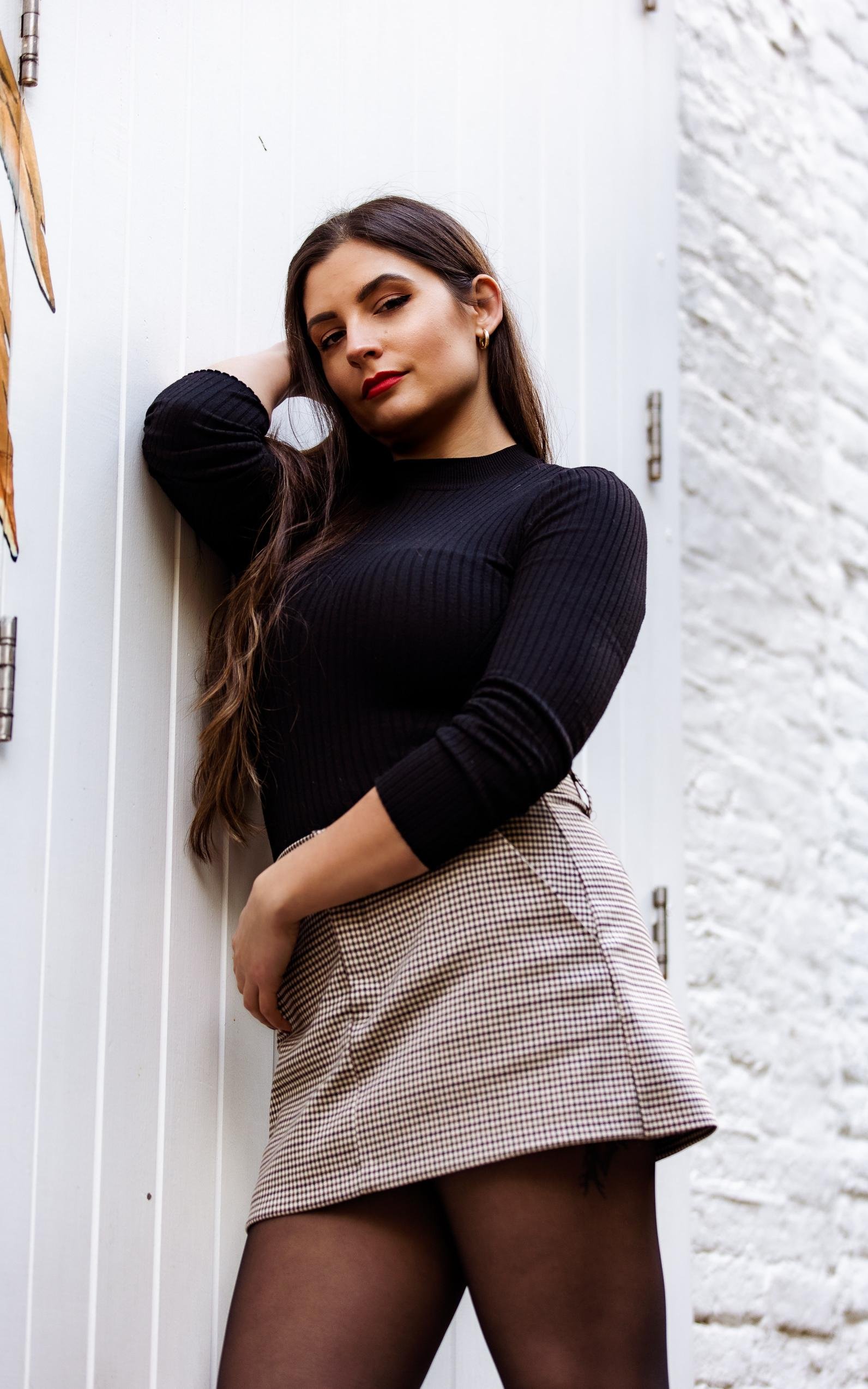 Confident woman in a beige plaid high-waisted skirt and black top posing by a white wall
