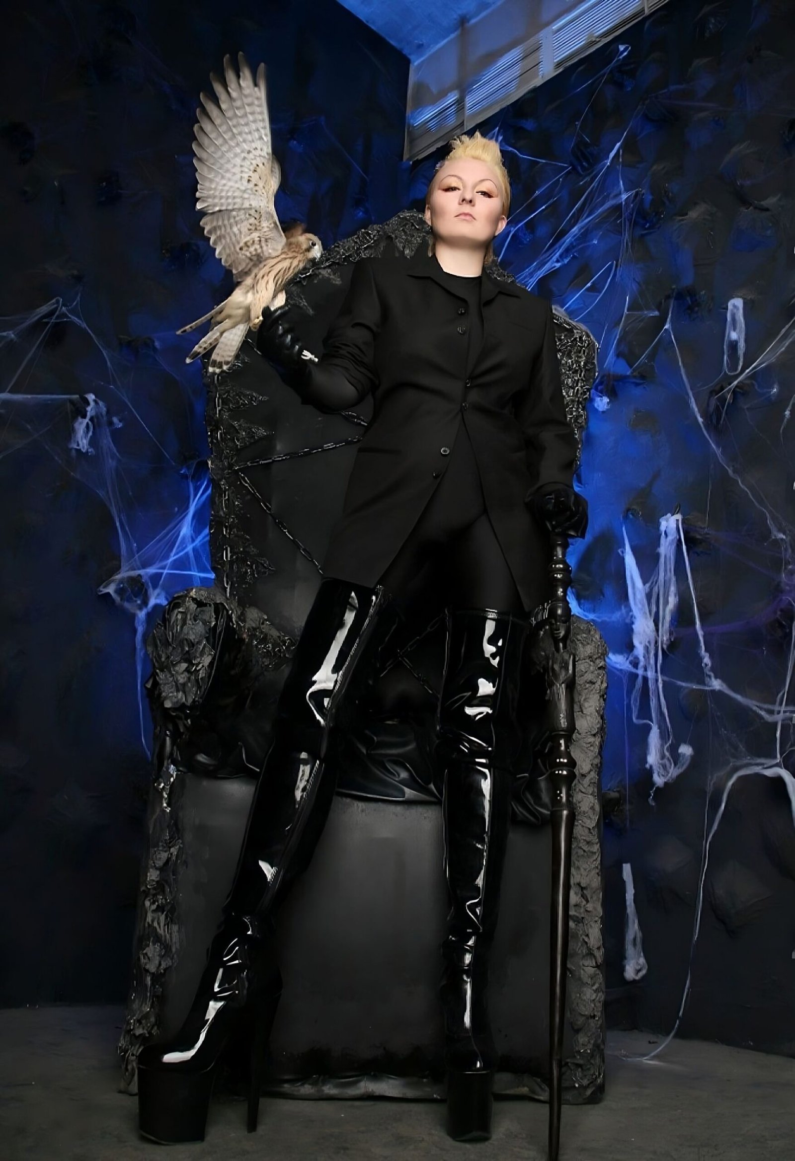 Dominant woman in black latex thigh-high boots and gothic throne holding a hawk