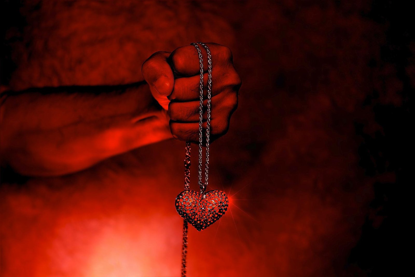 Hand gripping a chain with a blood-red heart pendant in dramatic lighting
