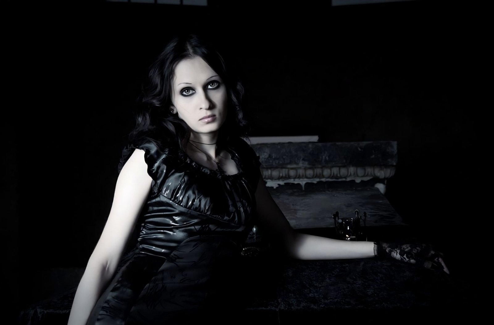 Gothic woman in black latex dress with dark eye makeup in a shadowy room