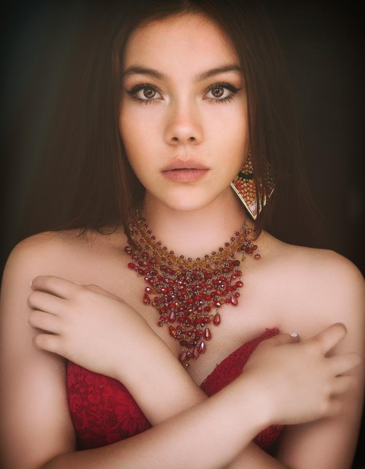 Woman in red lace corset and ornate gemstone necklace staring confidently into the camera 
