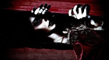 Close-up of vampire woman with dark makeup gripping the edge of a coffin lid
