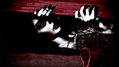 Close-up of vampire woman with dark makeup gripping the edge of a coffin lid