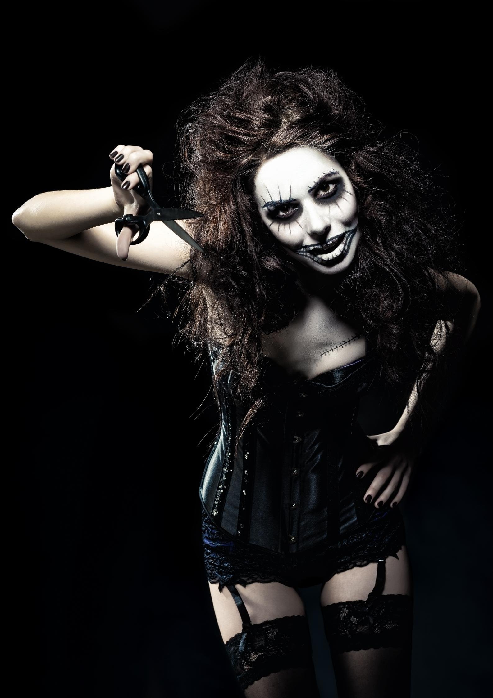Gothic woman in corset and lace stockings with skull face paint, holding scissors with a wild expression.