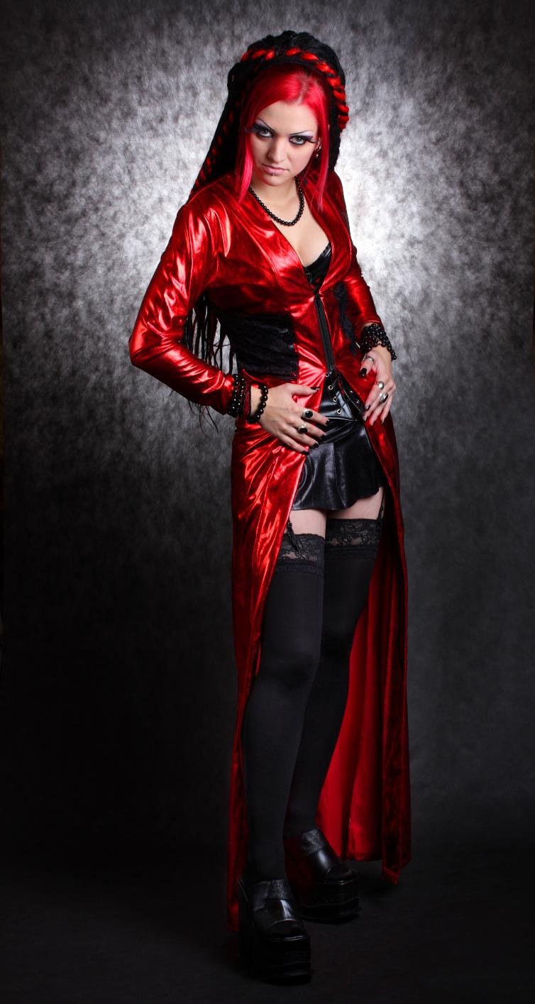 Gothic woman with long red and black braided hair wearing a red PVC jacket, black mini skirt, and thigh-high stockings, posing against a dark textured background.