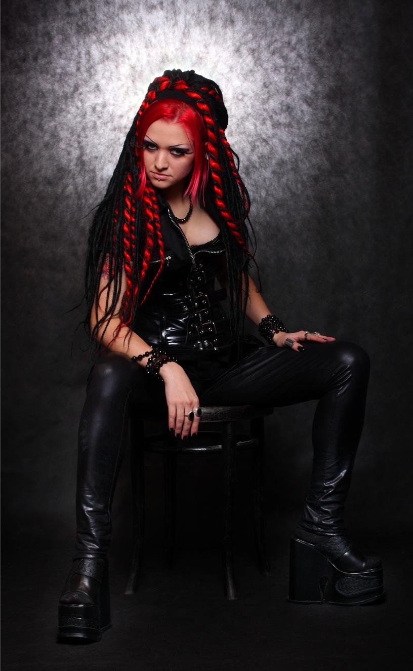 Gothic woman with red and black dreadlocks sitting on a wooden chair, dressed in a black leather corset and tight pants, exuding a dark and intense aura.
