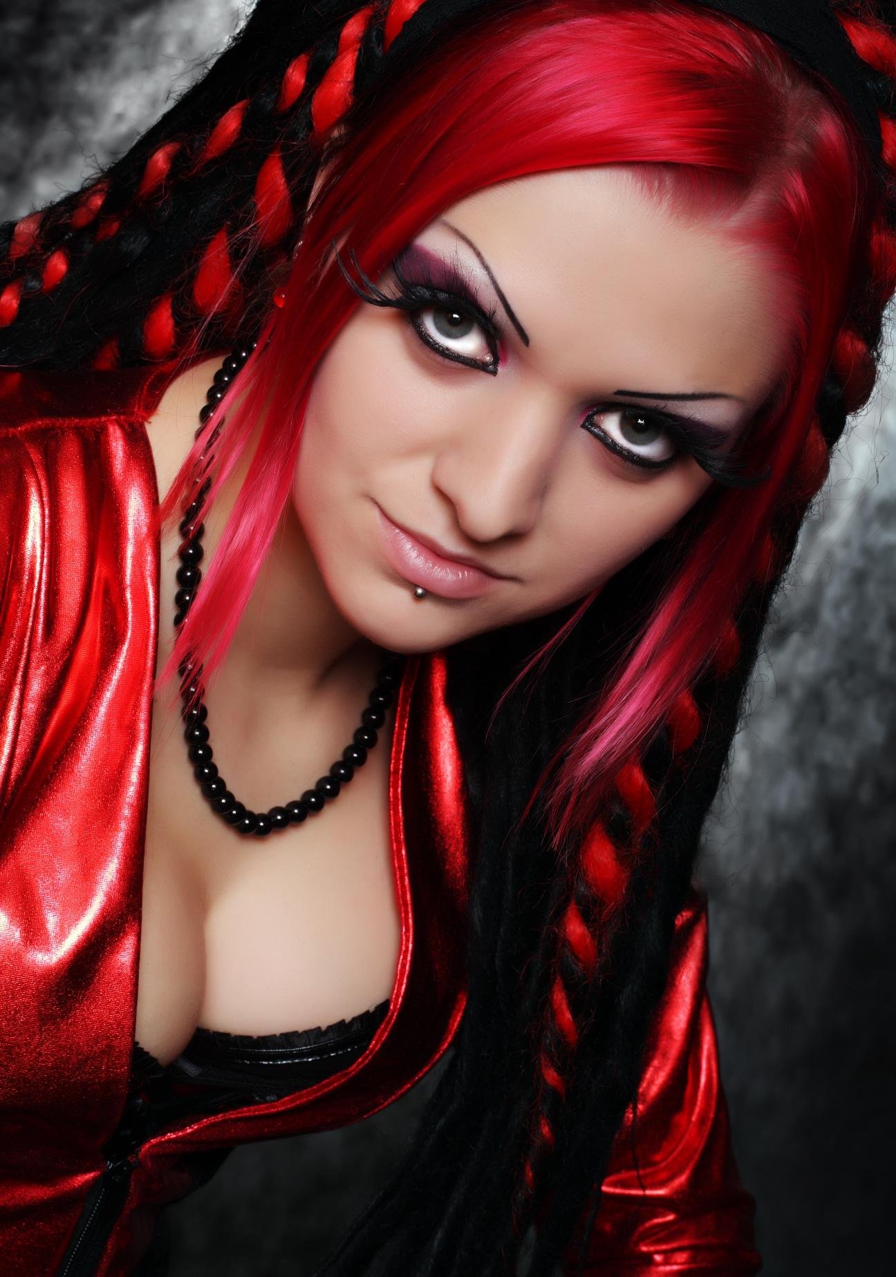 Close-up portrait of a gothic woman with striking red and black braids, intense eye makeup, and a deep neckline in a red PVC jacket.