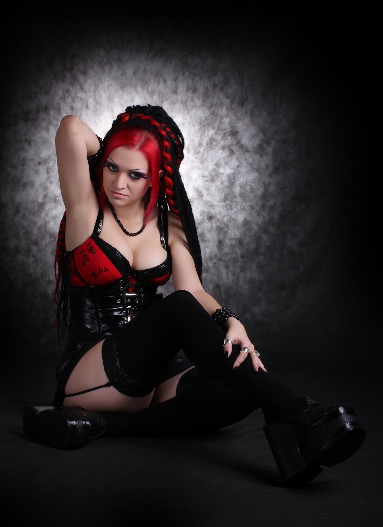Gothic woman sitting on the floor in a black corset with red lace accents, thigh-high stockings, and black platform boots, striking a fierce pose.