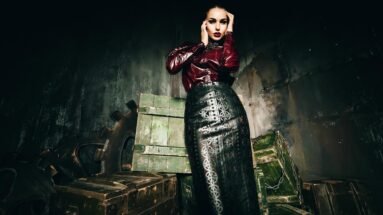 Woman in burgundy latex top and black leather pencil skirt standing against industrial crates