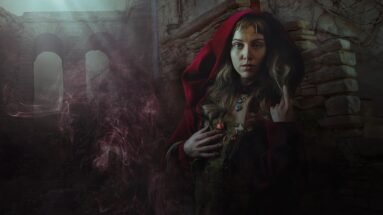 Woman in red cloak standing in gothic ruins with mist, flowers on dress, and a fierce stare.