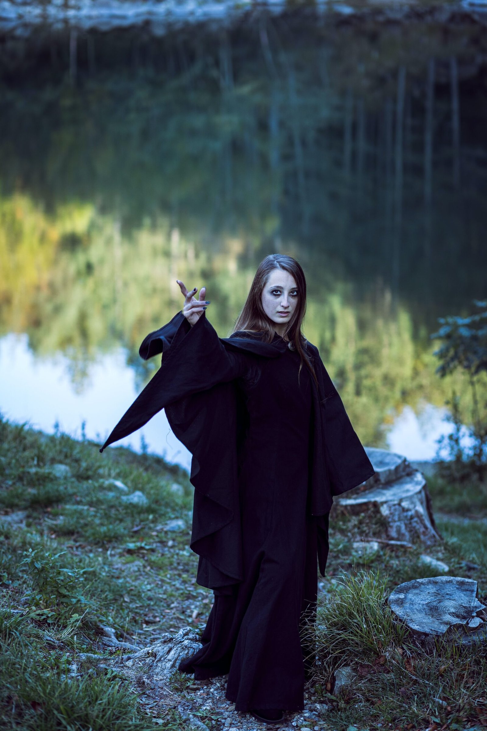 Gothic woman in flowing black cloak casting a spell beside a lake in a forest setting.