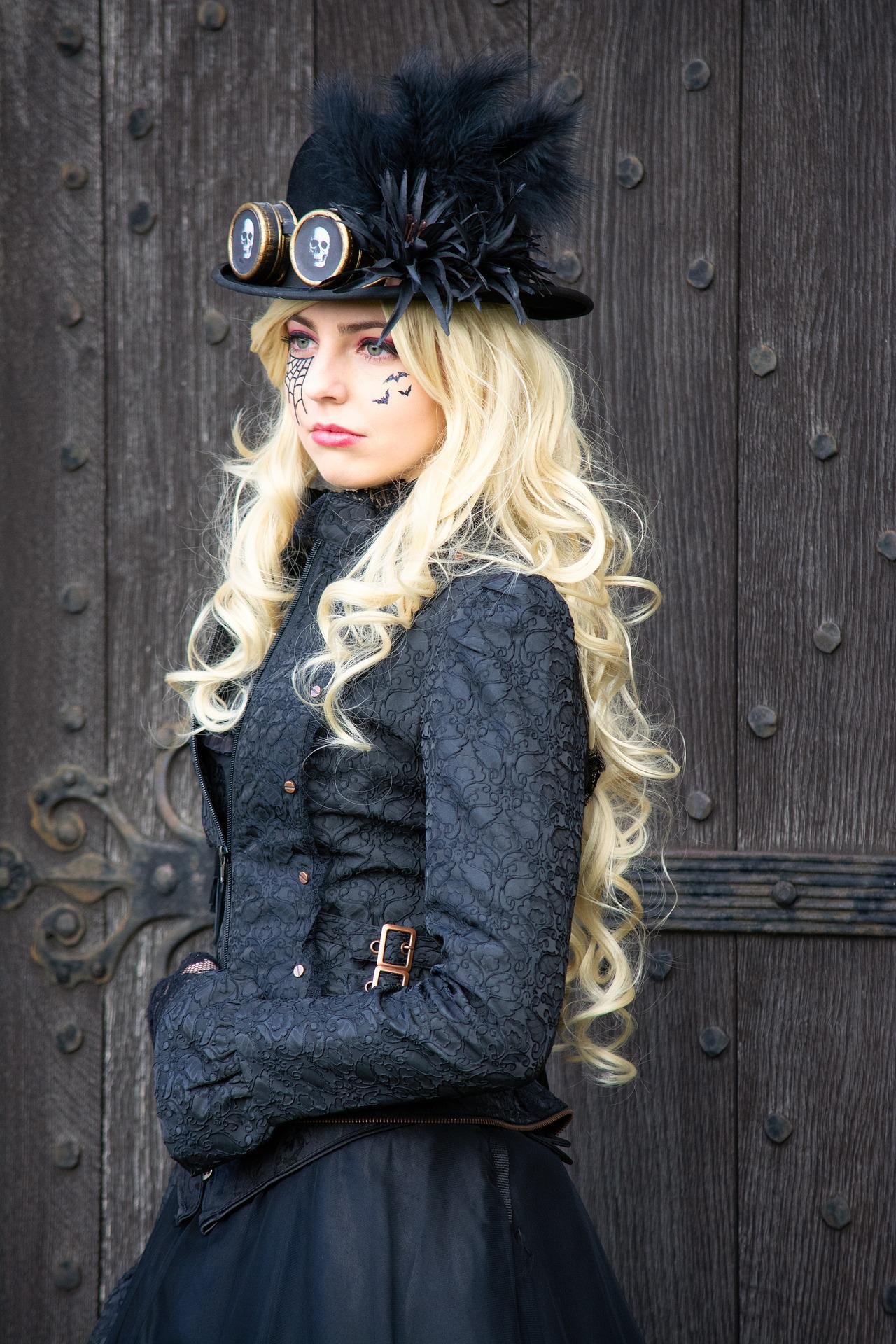 Steampunk goth woman in skull goggles, top hat, and spiderweb face paint standing by a wooden door.