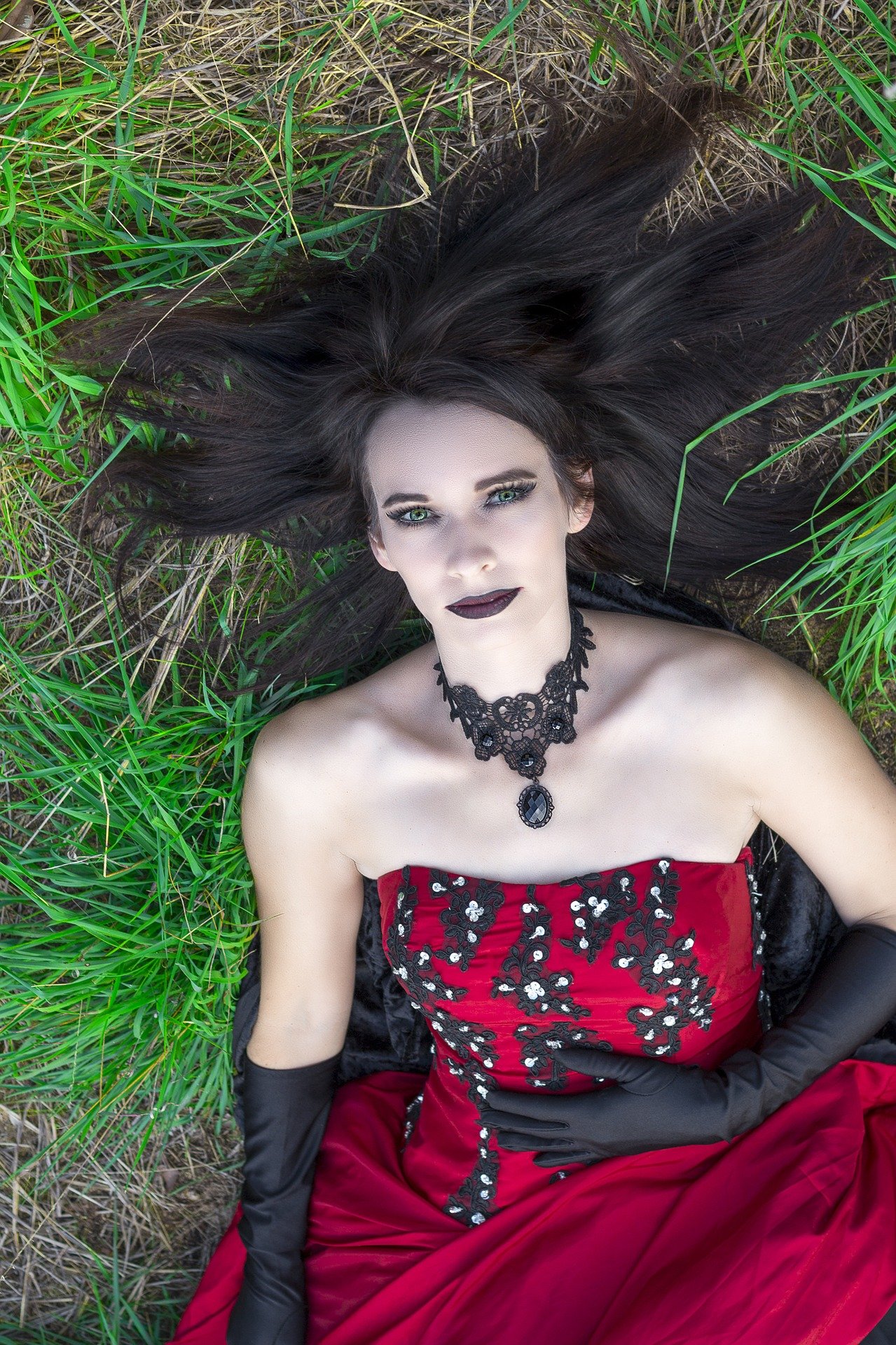 Gothic woman in red dress with lace choker lying on grass, dark makeup and long black hair spread around.