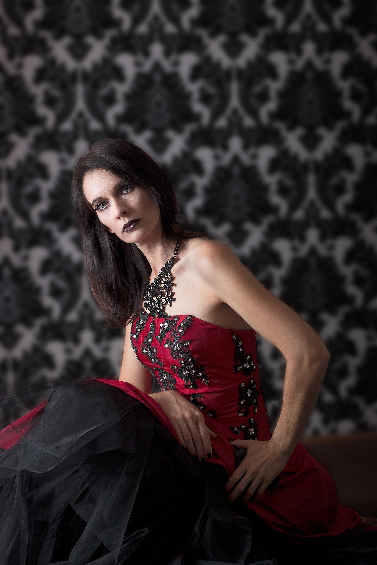 Woman in gothic red corset dress sitting indoors with black wallpaper, dark lipstick and straight dark hair.