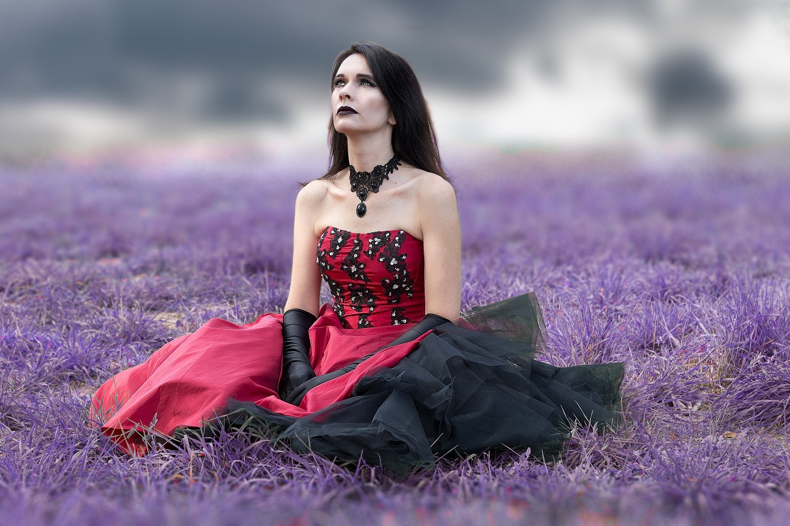 Gothic woman in red and black gown sitting in a purple grass field, looking up with a distant expression.