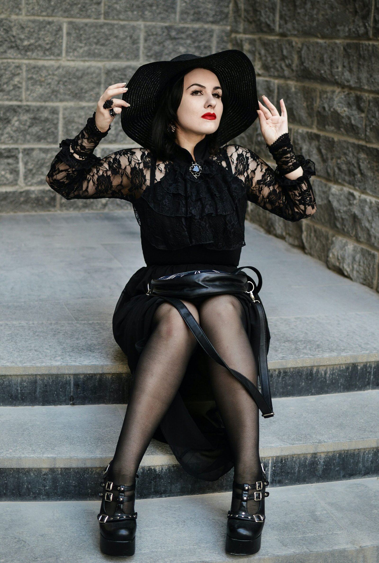 Goth woman in black lace outfit and hat sitting on stairs, wearing red lipstick and platform heels. 