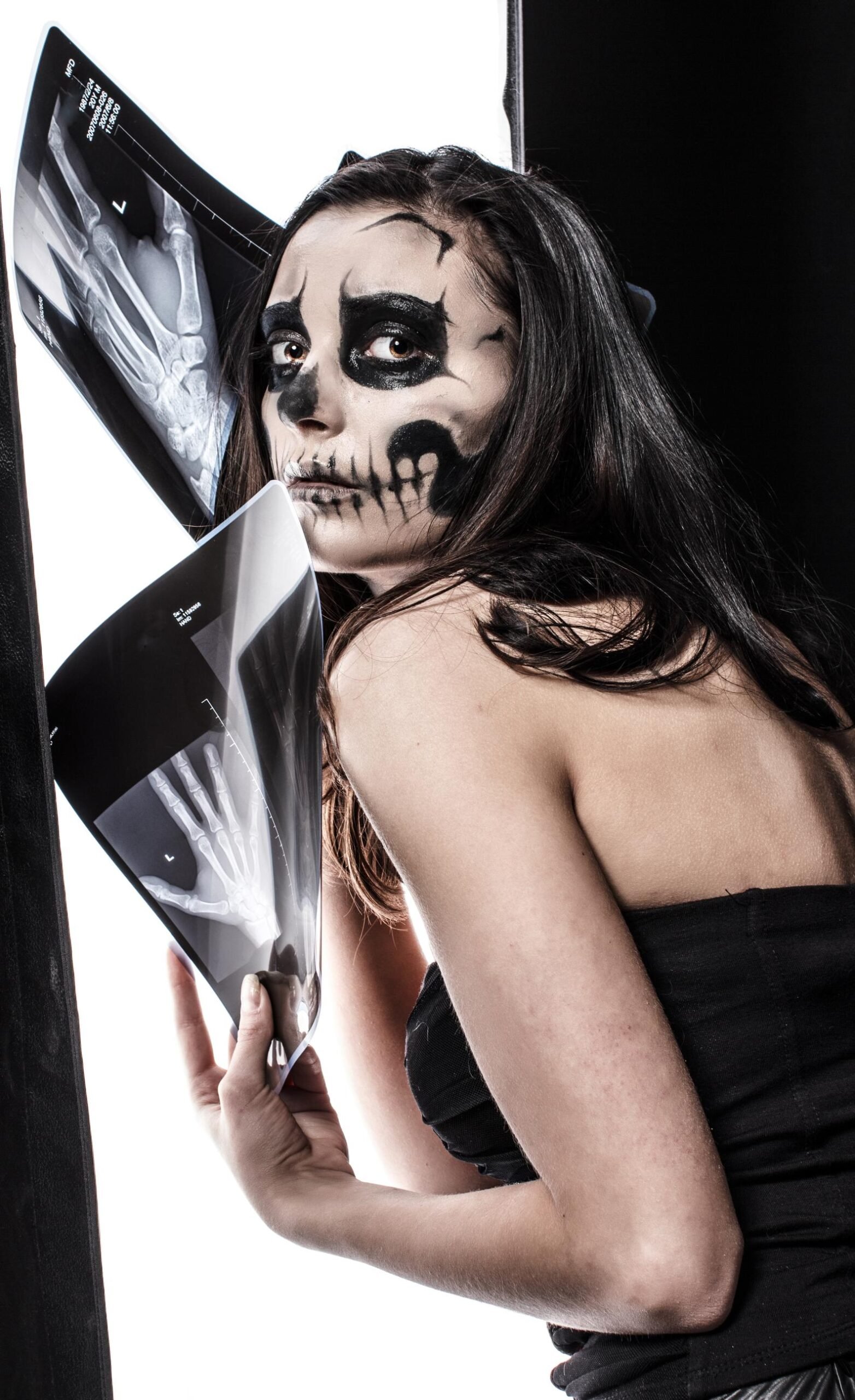 Woman with skeletal black face paint holding x-ray images, wearing a black strapless top against a high-contrast white and black studio background.